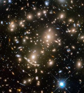 A packed field of galaxies and curved blue streaks and arcs. Large grouping of galaxies warped around what appears to be a giant lens. Each galaxy appears as a glob of light. Bright blue-white foreground star at bottom right.